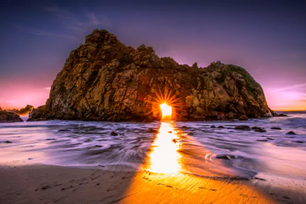 HD wallpaper of a stunning sunset over the ocean, with sunlight streaming through a natural rock arch on a sandy beach. The sun's rays create a beautiful golden path on the water and sand.