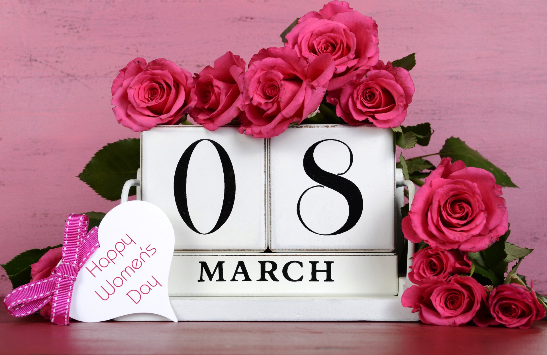 A beautiful arrangement of pink roses surrounds a calendar displaying 08 March with a heart-shaped tag that reads Happy Women's Day, capturing a festive and celebratory atmosphere.
