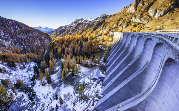 HD PC desktop wallpaper and background: man-made concrete dam curving through a snowy mountain winter landscape, golden trees and a clear blue sky.