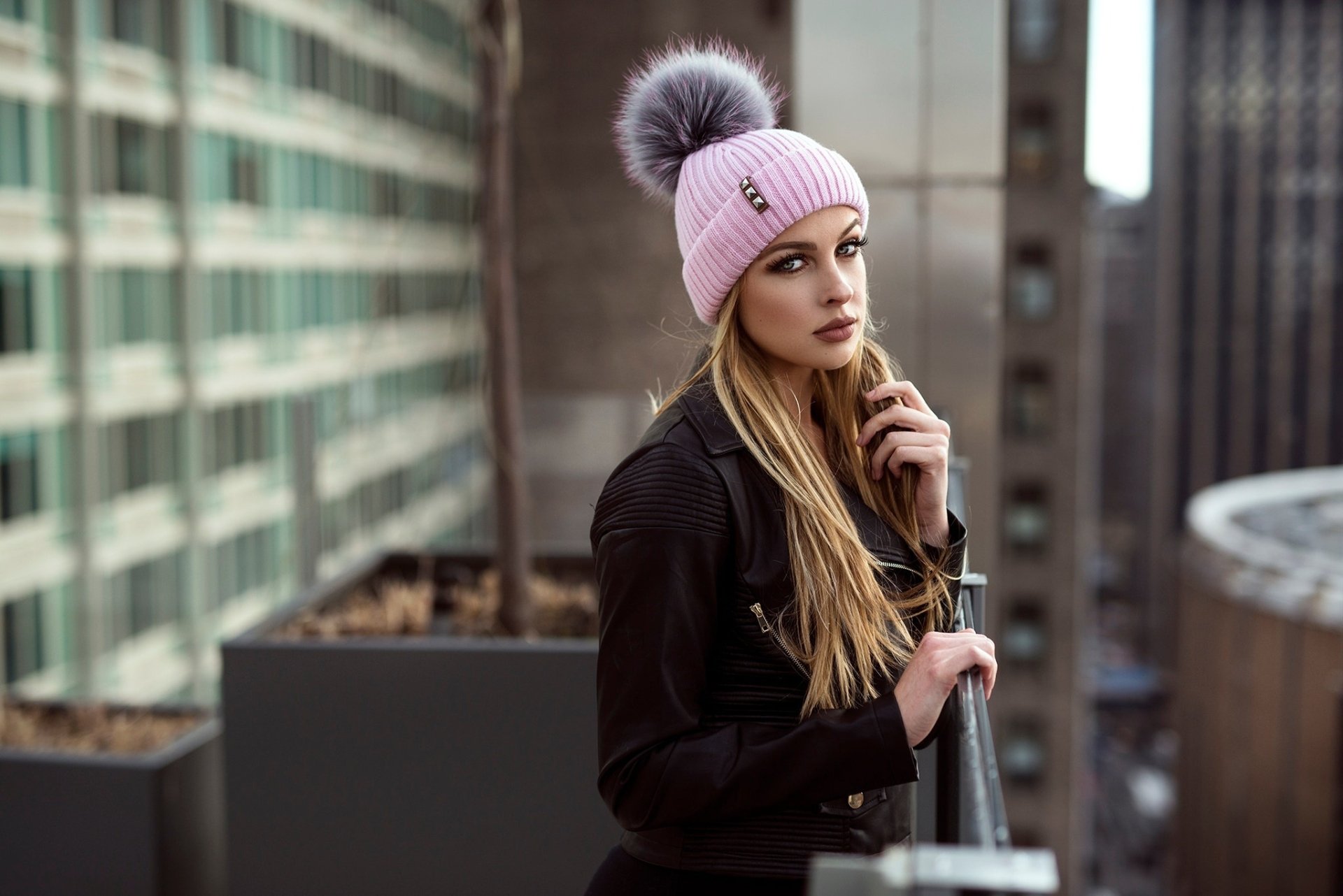 HD PC desktop wallpaper of a blonde woman model in a pink pom‑pom beanie, leaning on a rooftop railing with a blurred urban background and shallow depth of field.