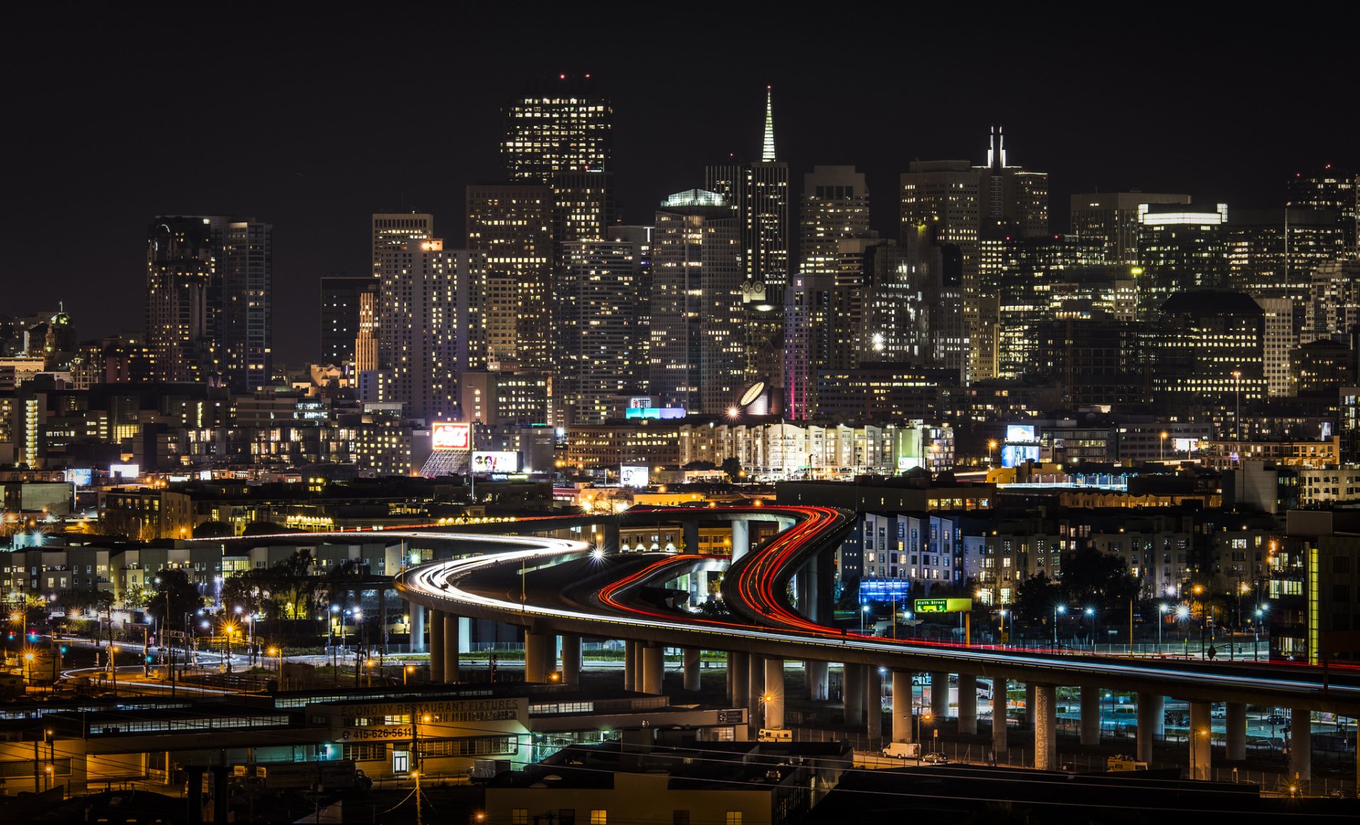 Nighttime time-lapse view of San Francisco’s illuminated skyline with skyscrapers and curving highways glowing against the dark sky in this HD cityscape wallpaper.
