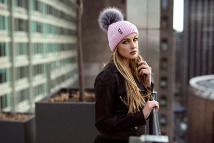 HD PC desktop wallpaper of a blonde woman model in a pink pom‑pom beanie, leaning on a rooftop railing with a blurred urban background and shallow depth of field.