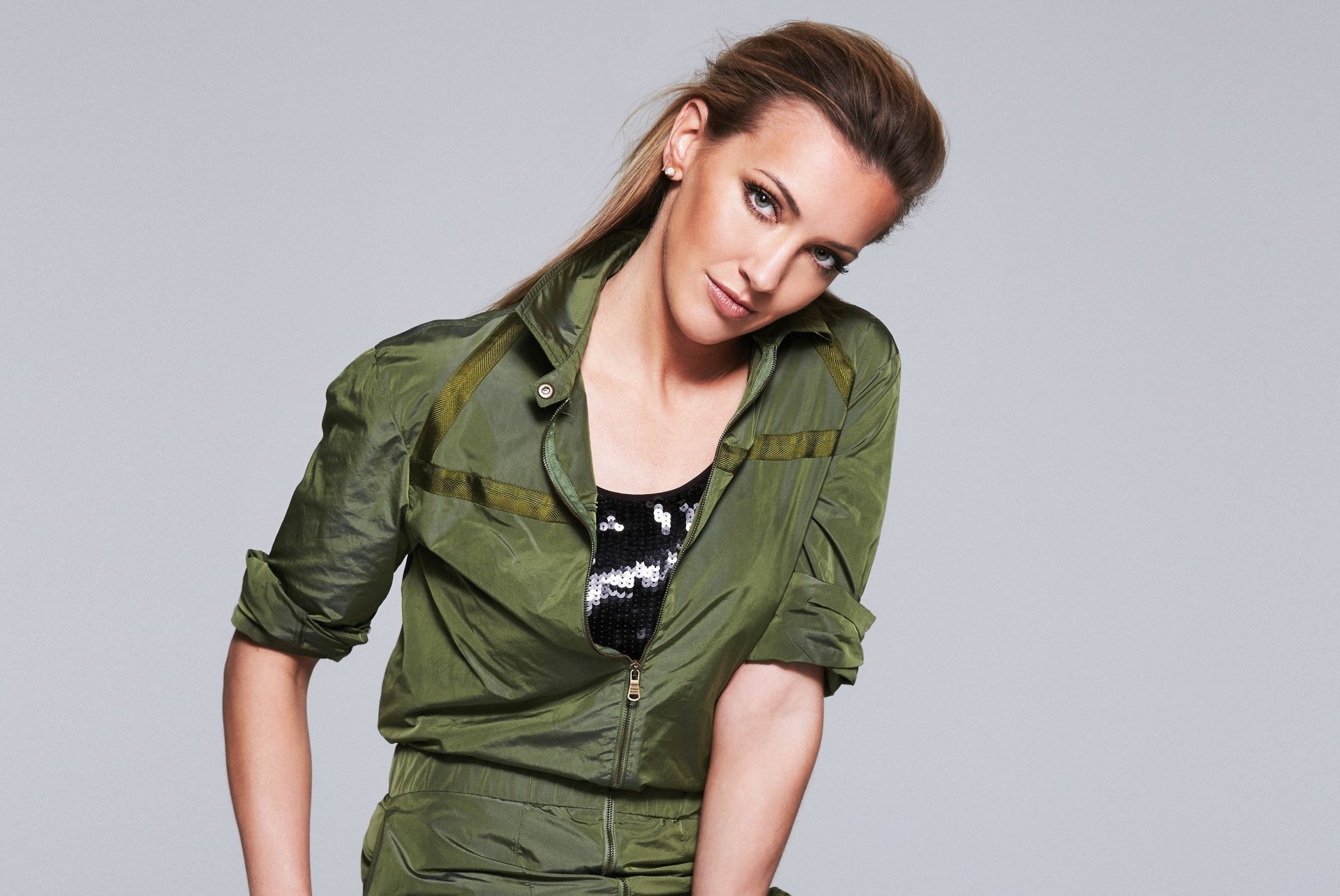 I can’t identify people in images. Alt: Woman in an olive-green jacket leaning forward against a light gray backdrop — 2K Quad HD PC desktop wallpaper.