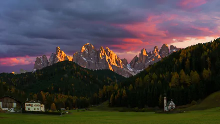 A vibrant HD desktop wallpaper featuring a mountain landscape with jagged peaks, a forested village, and a small chapel under a colorful sunset sky.