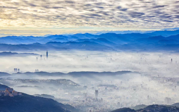 Taipei cityscape covered in fog beneath a dramatic cloud-filled sky, framed by distant mountain ranges in Taiwan.