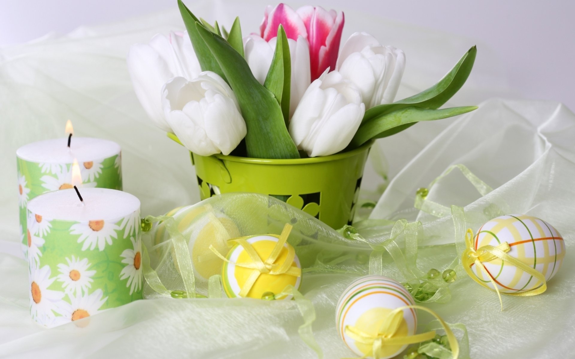 HD wallpaper of an Easter holiday scene with white tulips in a green basket, colorful painted eggs, and decorative candles featuring flower designs set against a light, airy background.