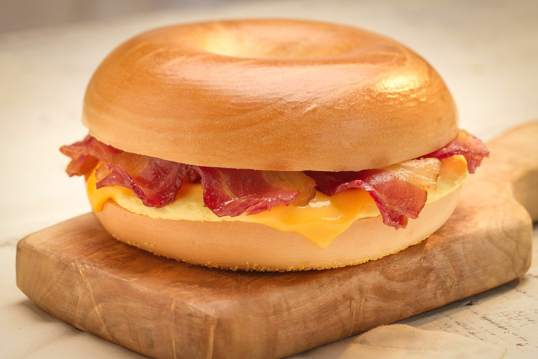 HD desktop wallpaper featuring a bacon and cheese sandwich with crispy bacon strips on a toasted bagel, served on a wooden cutting board.