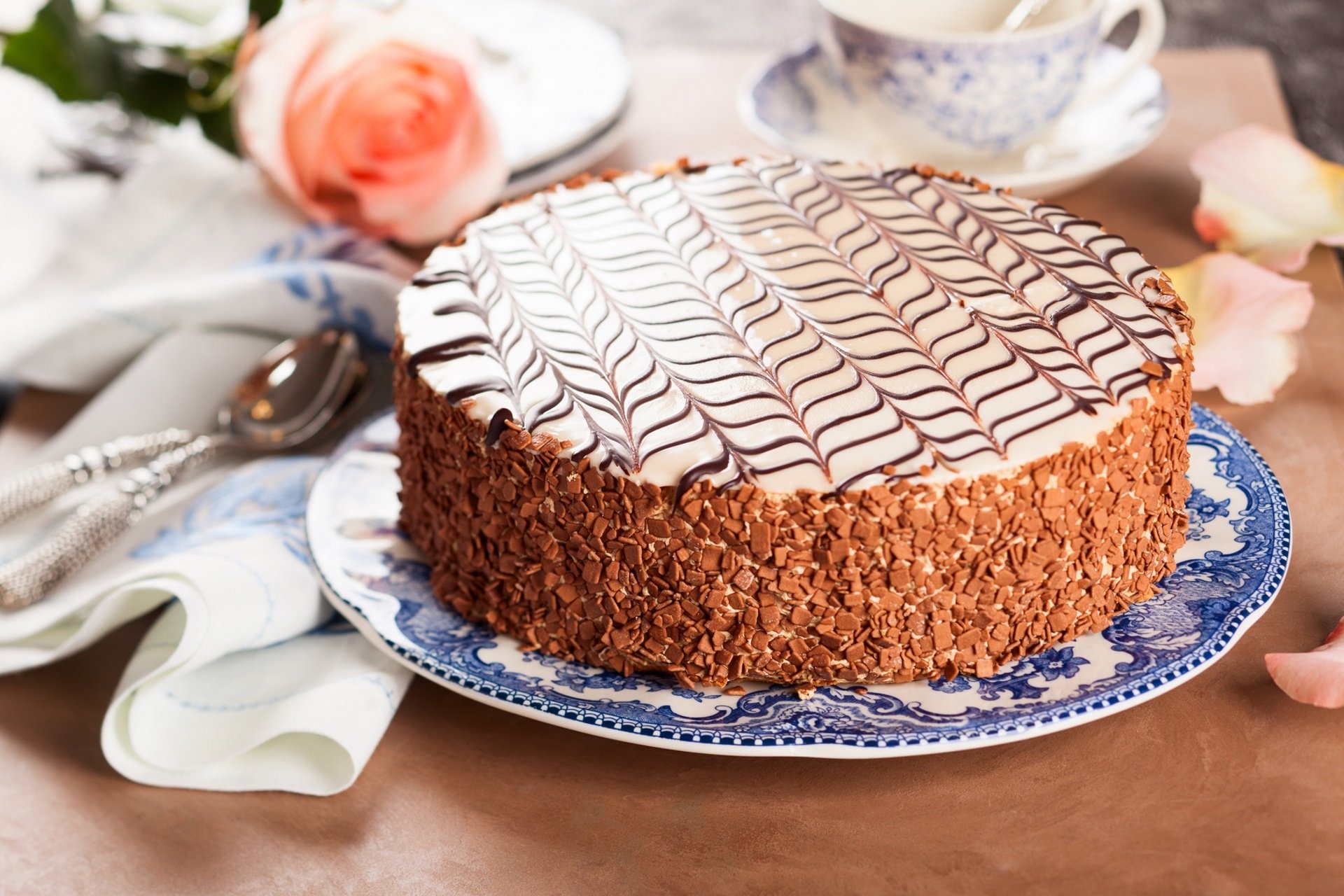 HD PC desktop wallpaper: pastry, food — close-up of a round chocolate cake with white zigzag icing and crunchy crumb sides on a blue floral plate, with a rose and teacup.