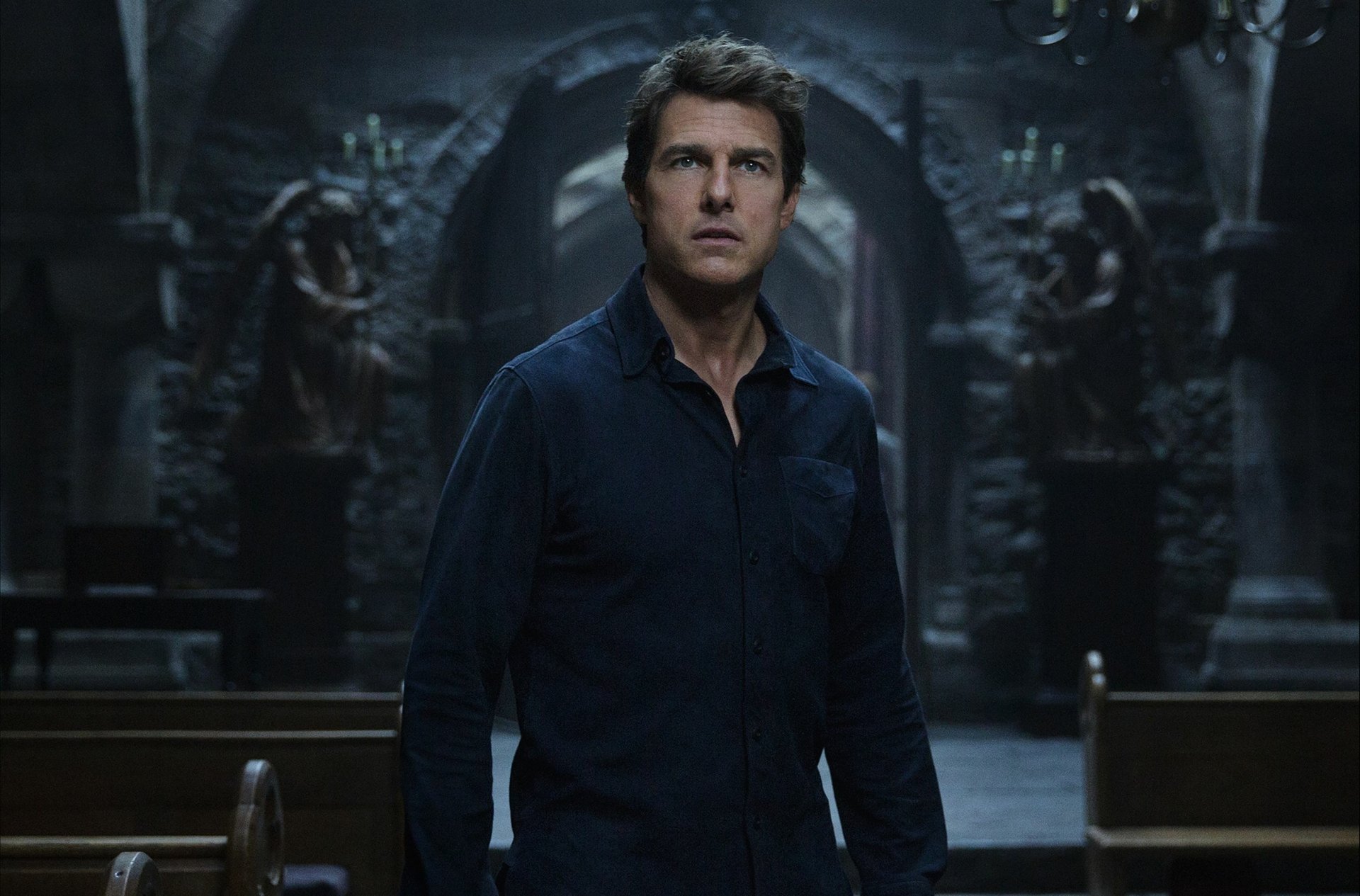 HD desktop wallpaper of Tom Cruise in a scene from the 2017 movie The Mummy. He stands in a dark, ornate room with stone statues in the background, wearing a dark shirt and looking intently forward.