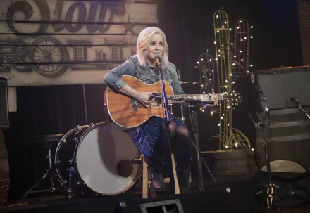 iZombie TV show wallpaper: lead character seated onstage playing an acoustic guitar amid drums and stage lights — 2K Quad HD PC desktop background.