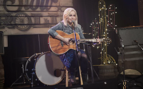 iZombie TV show wallpaper: lead character seated onstage playing an acoustic guitar amid drums and stage lights — 2K Quad HD PC desktop background.