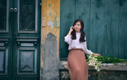 5K Ultra HD PC wallpaper: brunette woman model with twintails in a white blouse and rust pleated skirt, holding lilies while leaning against a weathered teal wooden wall.