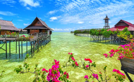Tropical Malaysian resort with overwater bungalows and huts on stilts above turquoise sea, vibrant pink flowers in foreground under a clear horizon sky.