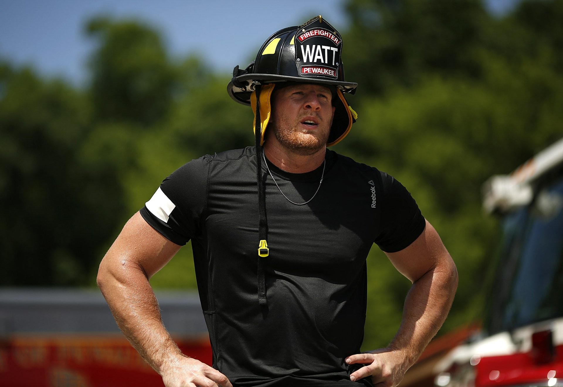 HD PC desktop wallpaper featuring J.J. Watt wearing a firefighter helmet and black athletic shirt, set outdoors with blurred greenery in the background, highlighting sports and strength.