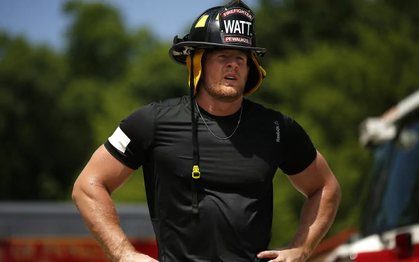 HD PC desktop wallpaper featuring J.J. Watt wearing a firefighter helmet and black athletic shirt, set outdoors with blurred greenery in the background, highlighting sports and strength.