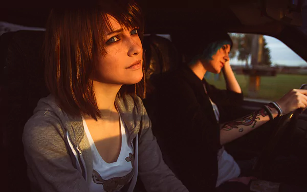 HD desktop wallpaper featuring women cosplaying Max Caulfield and Chloe Price from Life Is Strange, sitting in a car with sunlight casting a warm glow.