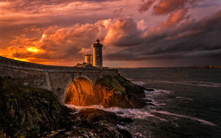 A dramatic sunset over the ocean, featuring a towering lighthouse on a rocky cliff connected by a stone arch bridge under a vibrant sky filled with colorful clouds. HD desktop wallpaper and background.