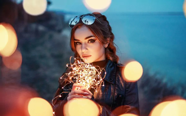 A brunette woman wearing sunglasses and holding fairy lights is captured in this HD portrait with a beautiful depth of field effect, making it an elegant desktop wallpaper and background.