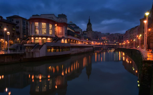 Night view of a canal in Bilbao, Spain, with reflections of architectural buildings and lights on the water, creating a serene atmosphere.