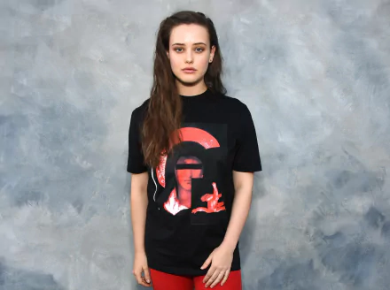 HD desktop wallpaper featuring Australian actress Katherine Langford, with brunette hair, standing against a textured backdrop, wearing a black graphic t-shirt.