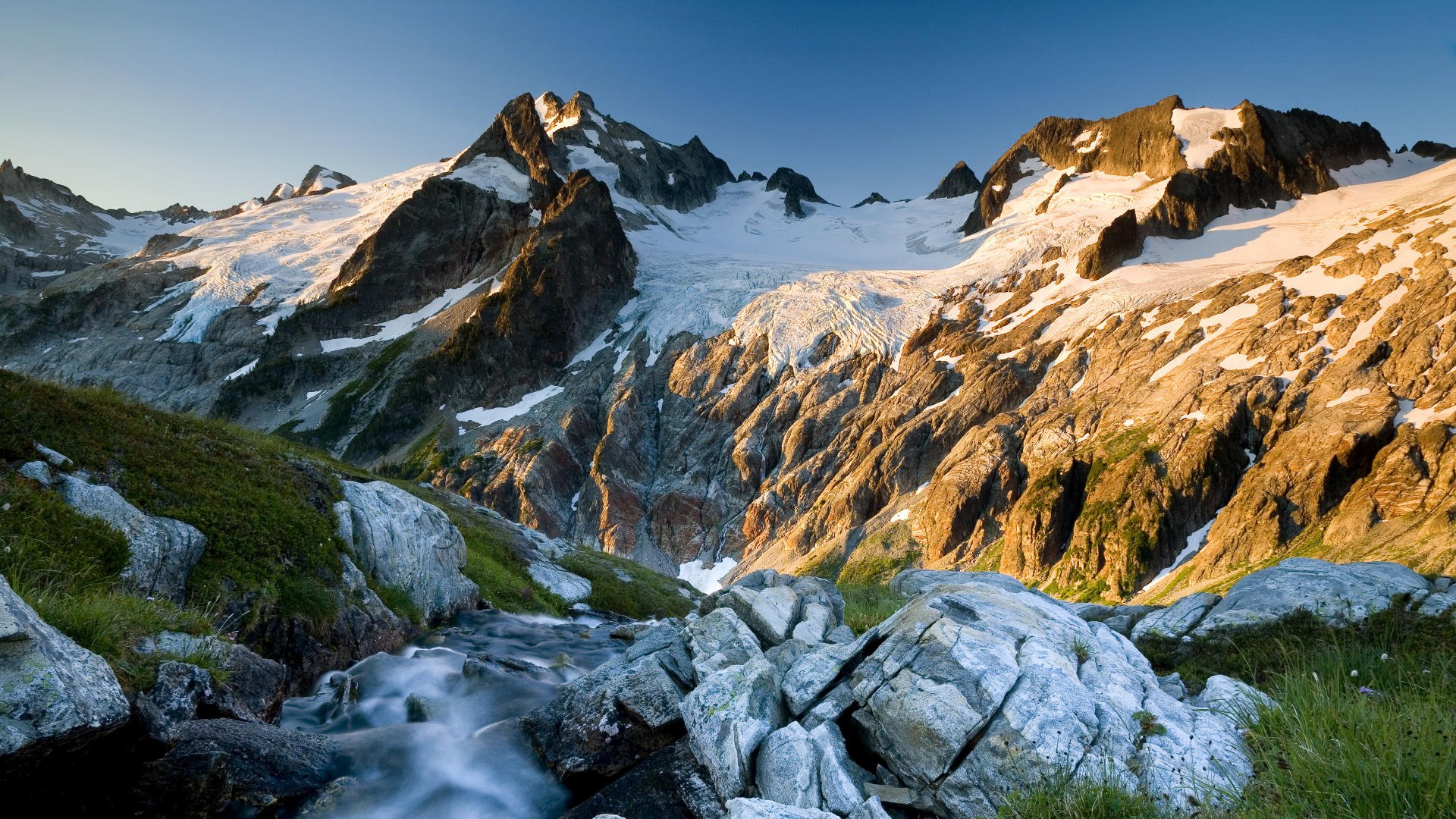 HD desktop wallpaper of a mountain landscape with a flowing stream, rocky foreground, and clear sky showcasing the beauty of nature.