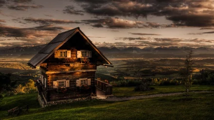 HDR image of a rustic cabin on a grassy hill during sunset, with a mountain range in the background, creating a breathtaking landscape. This HD desktop wallpaper exudes serene and natural beauty.