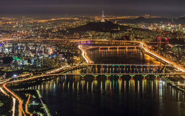 A stunning HD wallpaper of Seoul's night cityscape featuring illuminated bridges over a river, with the city lights creating a vibrant glow against the dark sky in South Korea.