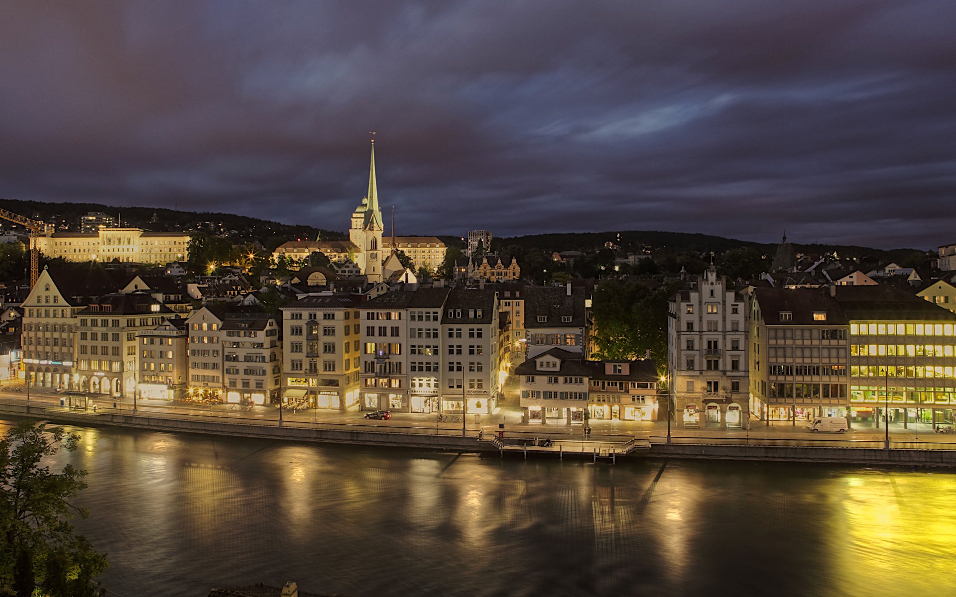 HD cityscape wallpaper capturing a man-made riverside with illuminated buildings under dramatic clouds at dusk.