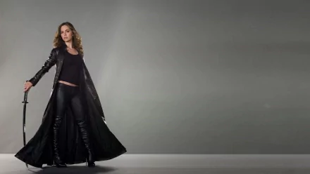 Eliza Dushku stands confidently in a sleek black outfit and a long coat, holding a sword against a minimalist gray background, creating a striking HD desktop wallpaper.