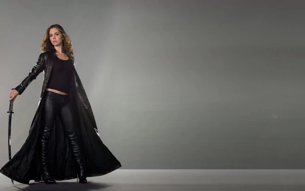 Eliza Dushku stands confidently in a sleek black outfit and a long coat, holding a sword against a minimalist gray background, creating a striking HD desktop wallpaper.
