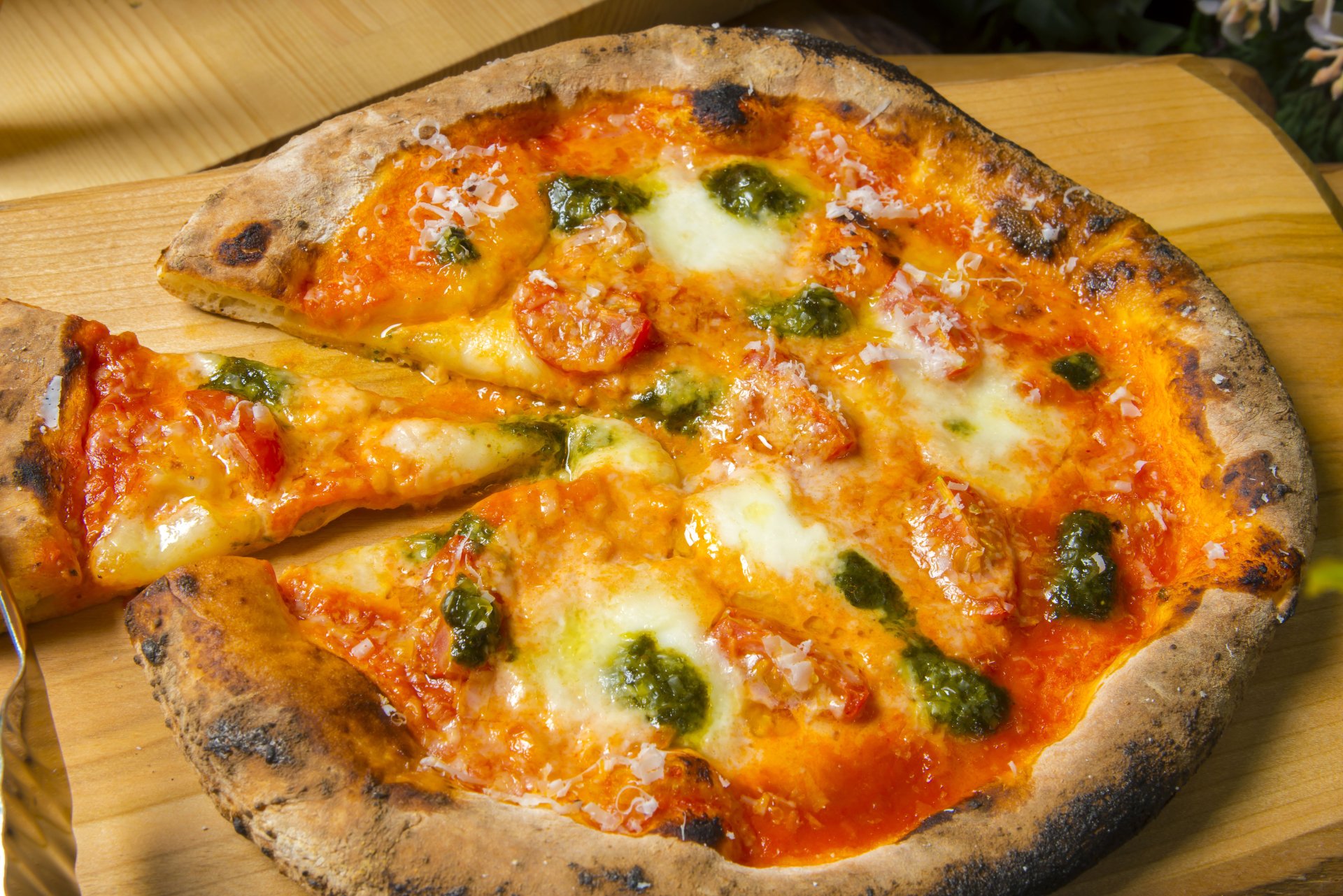 8K Ultra HD PC desktop wallpaper of food: a wood-fired pizza with charred crust, bubbling mozzarella, bright tomato sauce and basil pesto, one slice being lifted.