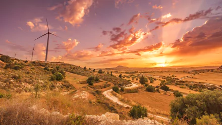 4K Ultra HD landscape photo of a Cyprus sunset: sunbeams through clouds over rolling hills, winding dirt track, scattered bushes and a wind turbine — PC desktop wallpaper.