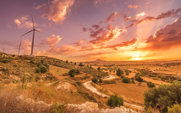 4K Ultra HD landscape photo of a Cyprus sunset: sunbeams through clouds over rolling hills, winding dirt track, scattered bushes and a wind turbine — PC desktop wallpaper.