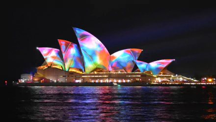 Colorful night view of the Sydney Opera House in Australia, illuminated with vibrant lights reflecting on the water. This HD desktop wallpaper captures the stunning architecture against a dark sky.
