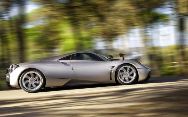 A stunning HD wallpaper of a silver Pagani Huayra supercar speeding on a road, set against a blurred background of lush green trees.