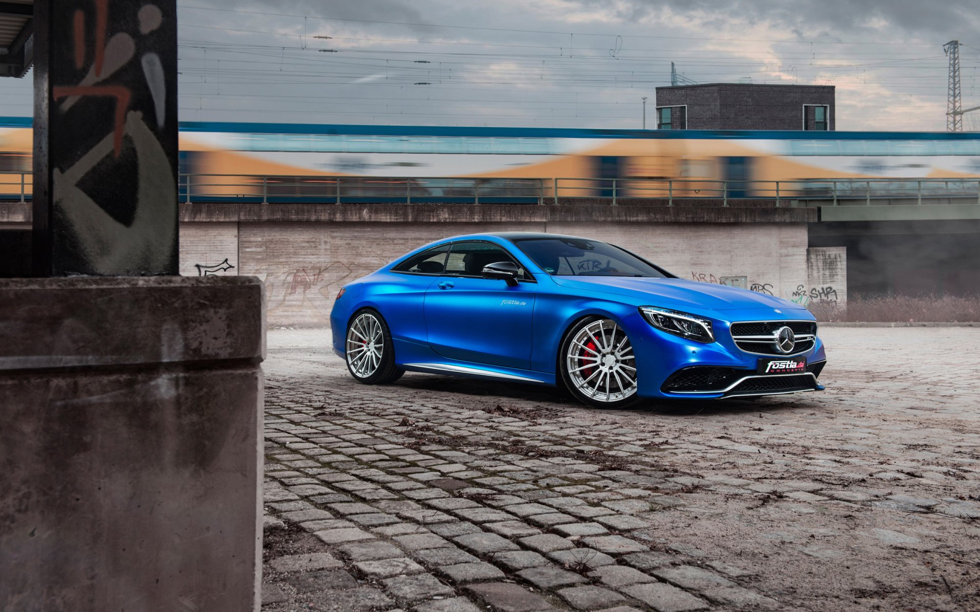 Blue Mercedes‑Benz S63 AMG on cobblestone quay beneath a cloudy sky, urban industrial backdrop — 2K Quad HD PC desktop wallpaper featuring the car and a passing train.