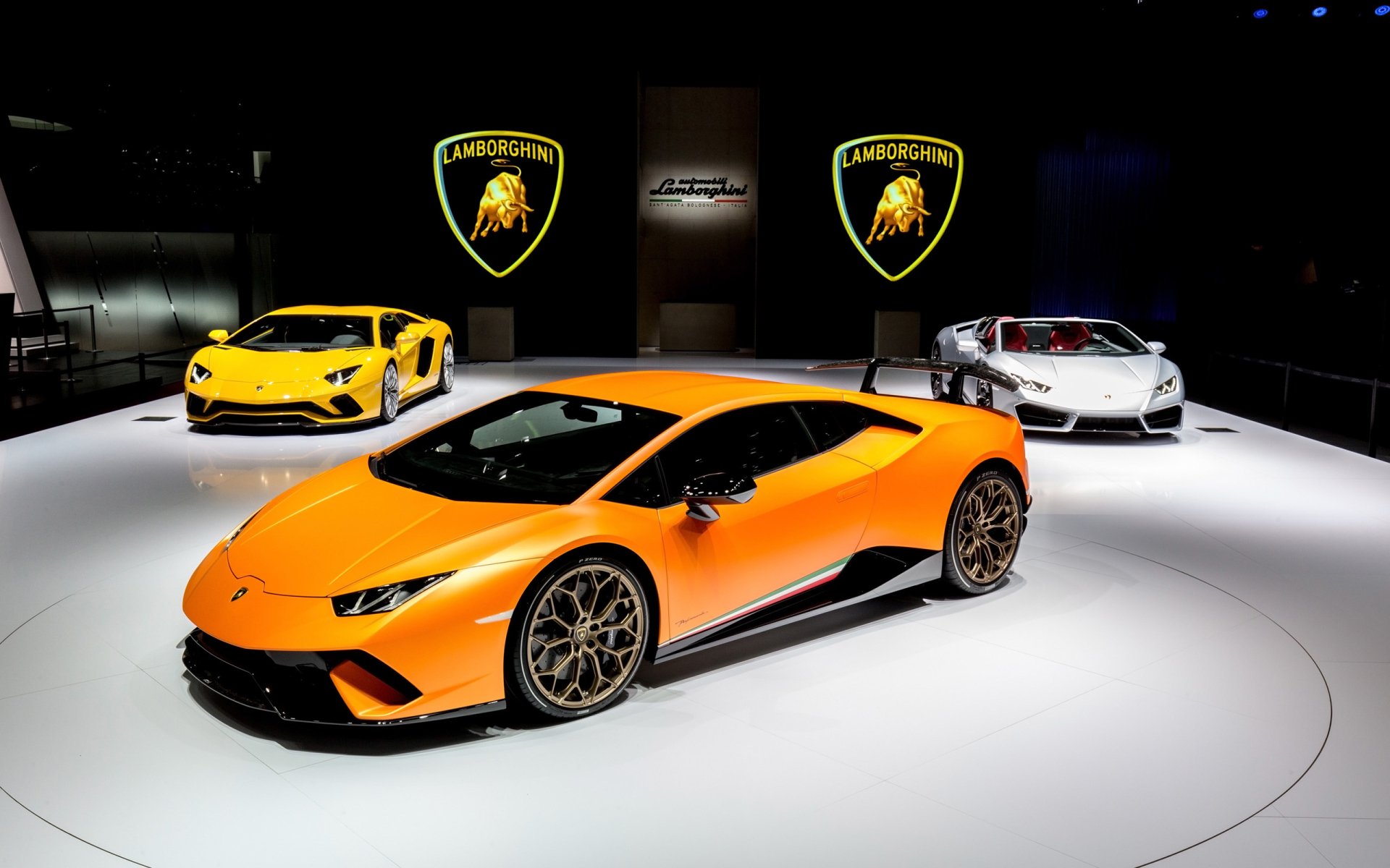 HD desktop wallpaper featuring three Lamborghini supercars, including an orange Lamborghini Huracan Performante, displayed in a sleek showroom with Lamborghini logos.