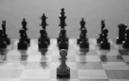 A black and white HD desktop wallpaper featuring a lone white pawn in front of a row of black chess pieces, set on a wooden checkerboard surface.