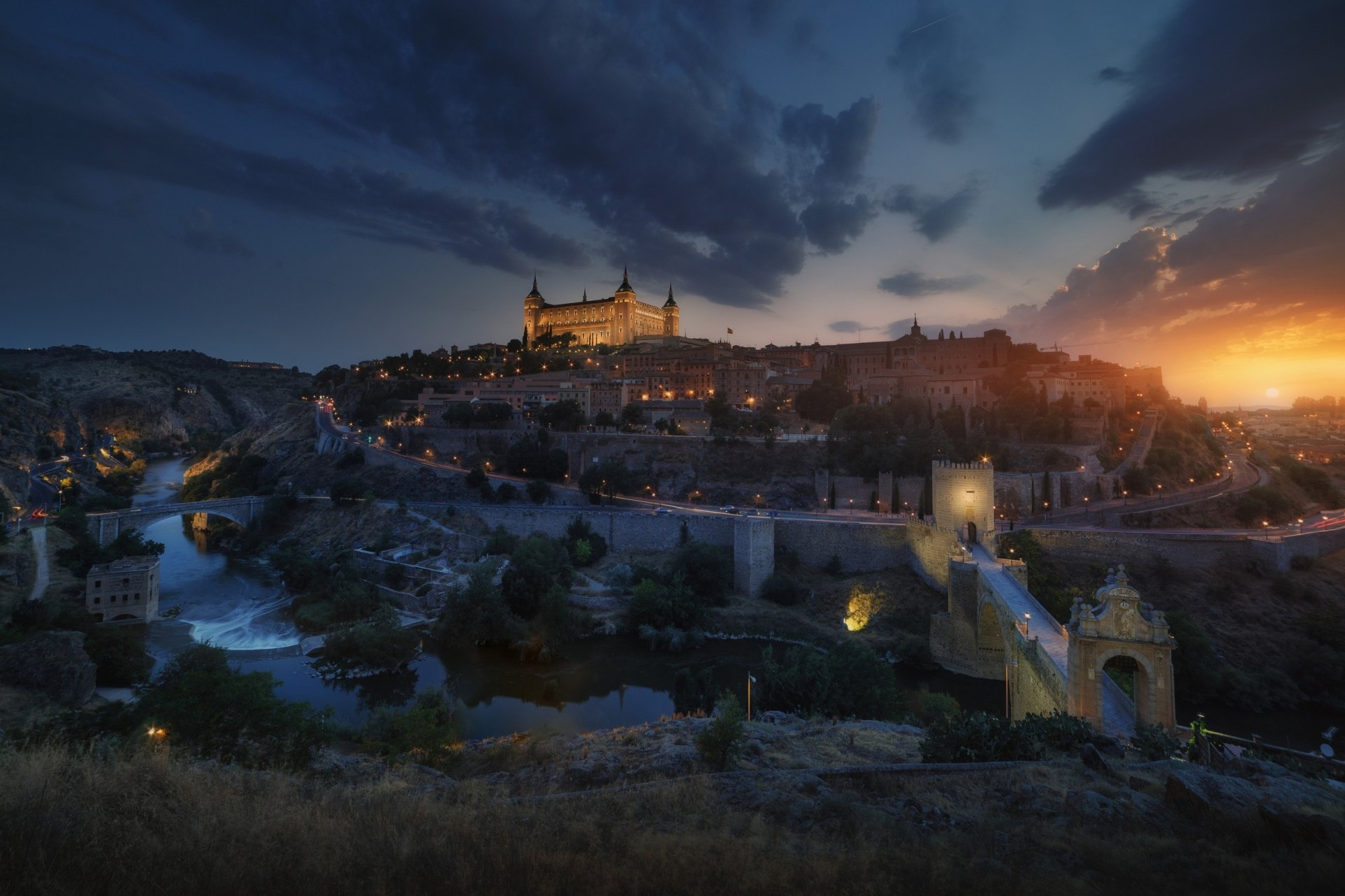 Night view of Toledo, Spain, showcasing historic buildings along the river with glowing lights illuminating the town under a dramatic sky.