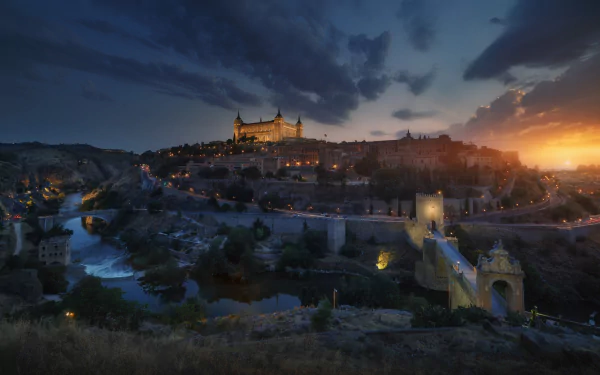 Night view of Toledo, Spain, showcasing historic buildings along the river with glowing lights illuminating the town under a dramatic sky.