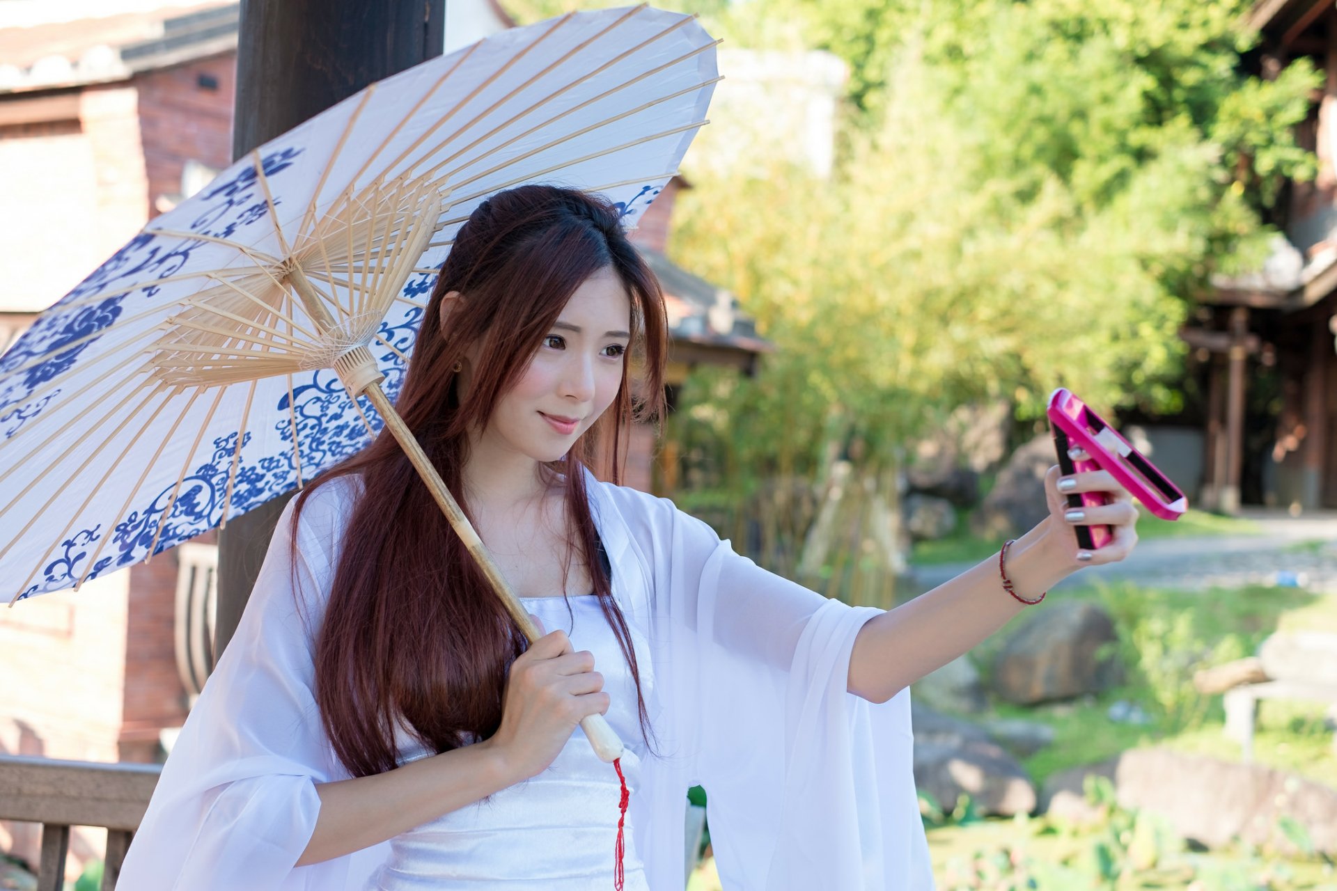 An Asian woman smiles while taking a selfie with her phone. She holds a decorative umbrella, standing outdoors on a sunny day. This HD image serves as a vibrant summer desktop wallpaper and background.
