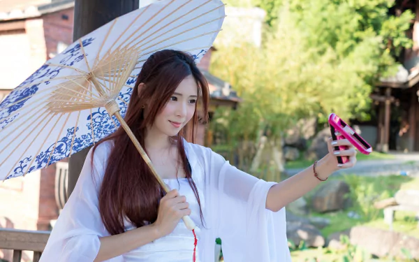 An Asian woman smiles while taking a selfie with her phone. She holds a decorative umbrella, standing outdoors on a sunny day. This HD image serves as a vibrant summer desktop wallpaper and background.