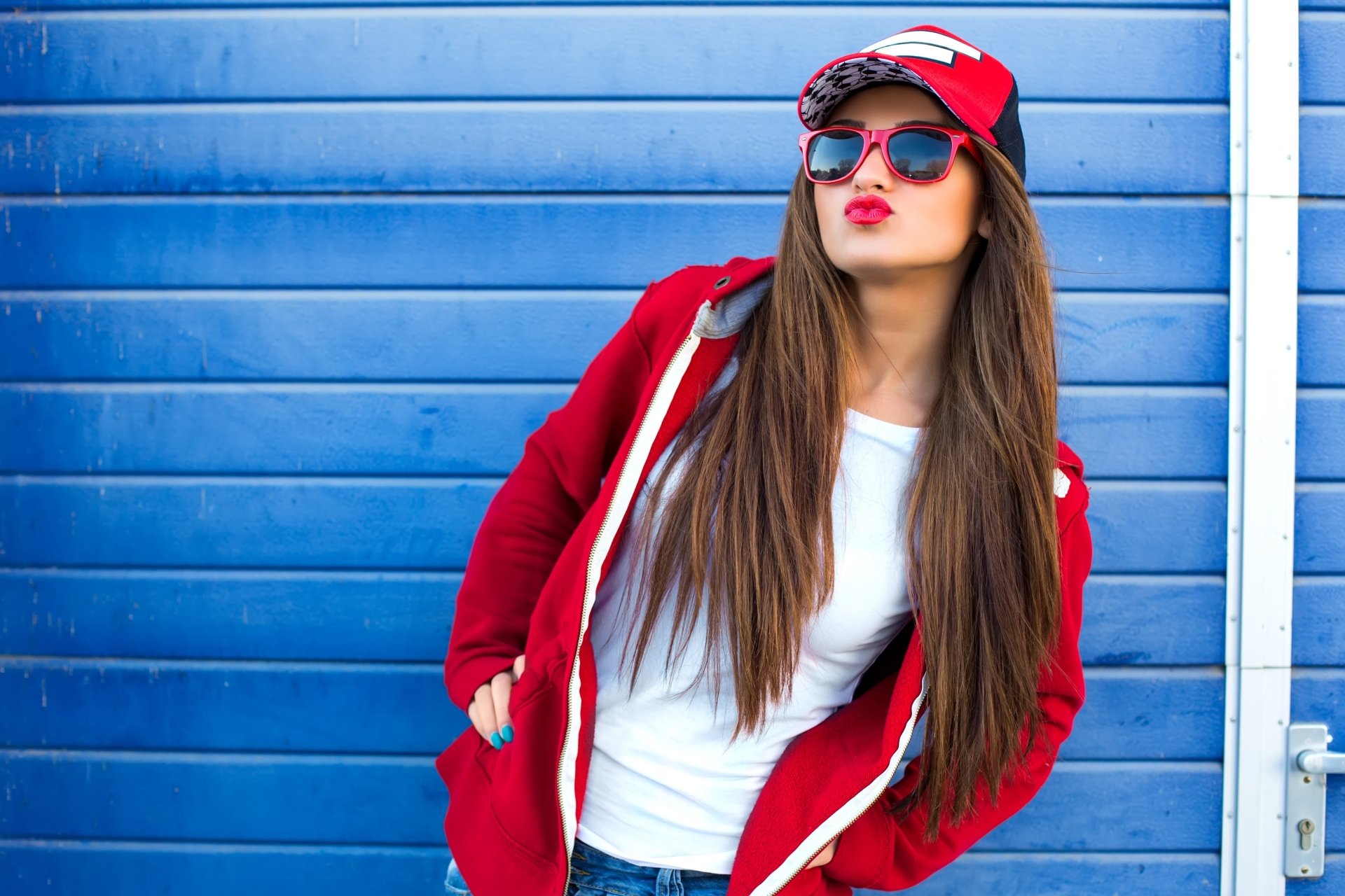Brunette model wearing a cap, sunglasses and red lipstick in a red hoodie, posing against a blue shutter — 4K Ultra HD PC desktop wallpaper and background.