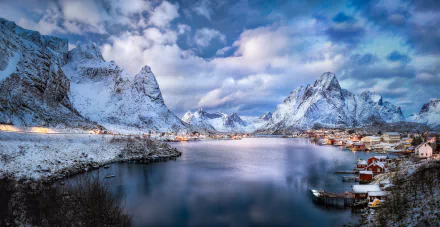 Winter landscape of Reine village in Lofoten, Norway, featuring snow-covered mountains, clouds, and a calm fjord under a vivid sky.