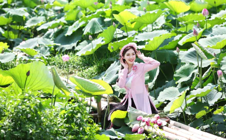 Brunette woman model smiling among lotus flowers and large green leaves, wearing a pink outfit on a bamboo walkway — HD desktop wallpaper background.