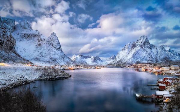 Winter landscape of Reine village in Lofoten, Norway, featuring snow-covered mountains, clouds, and a calm fjord under a vivid sky.