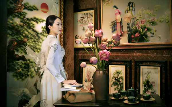 A Vietnamese woman in a traditional Ao Dai stands beside a table with a tea set, books, and a vase of lotus flowers, framed by floral paintings in a 4K Ultra HD desktop wallpaper.