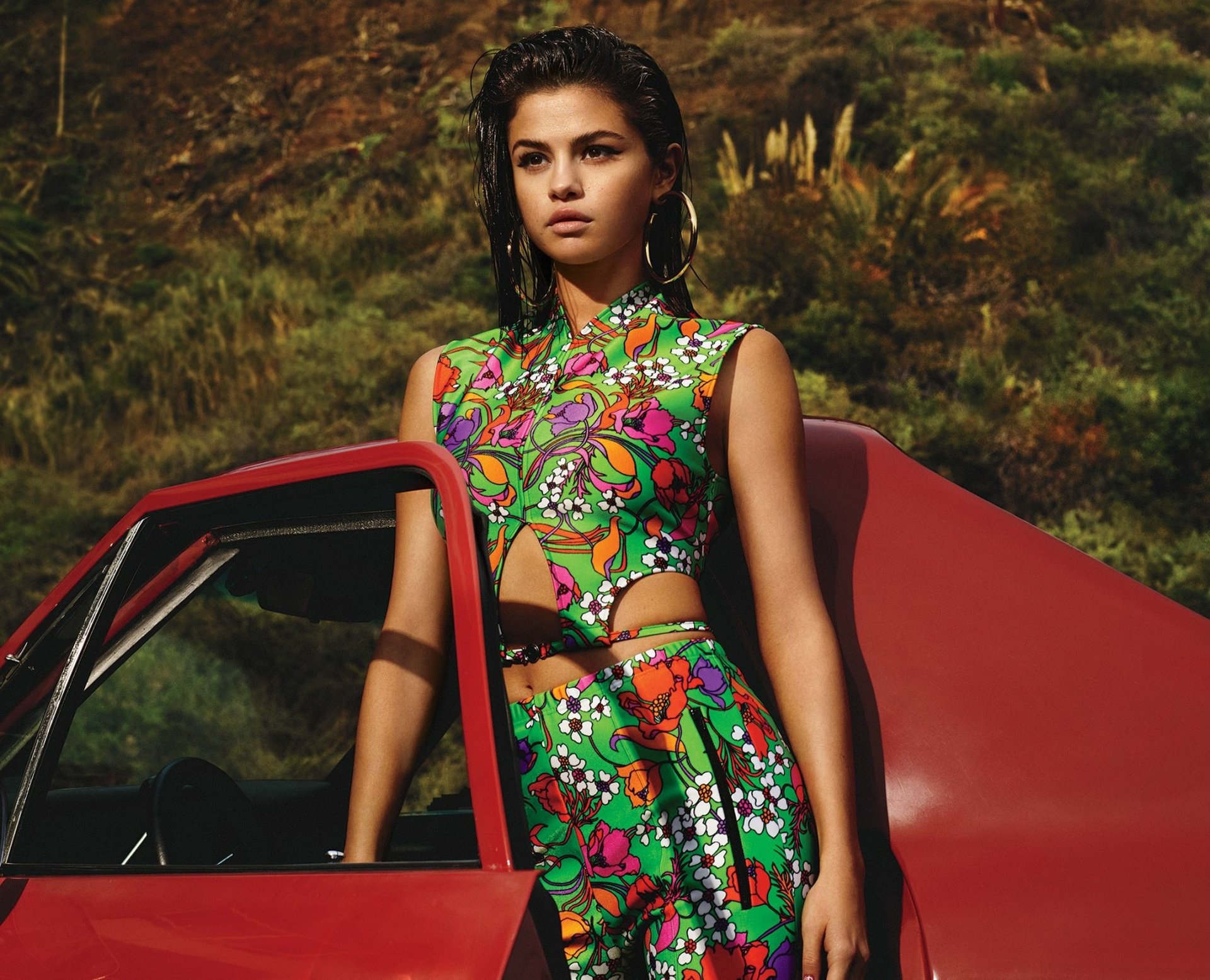 HD desktop wallpaper featuring American singer Selena Gomez, standing beside a red car in a colorful dress, with brunette hair, brown eyes, and wearing earrings.