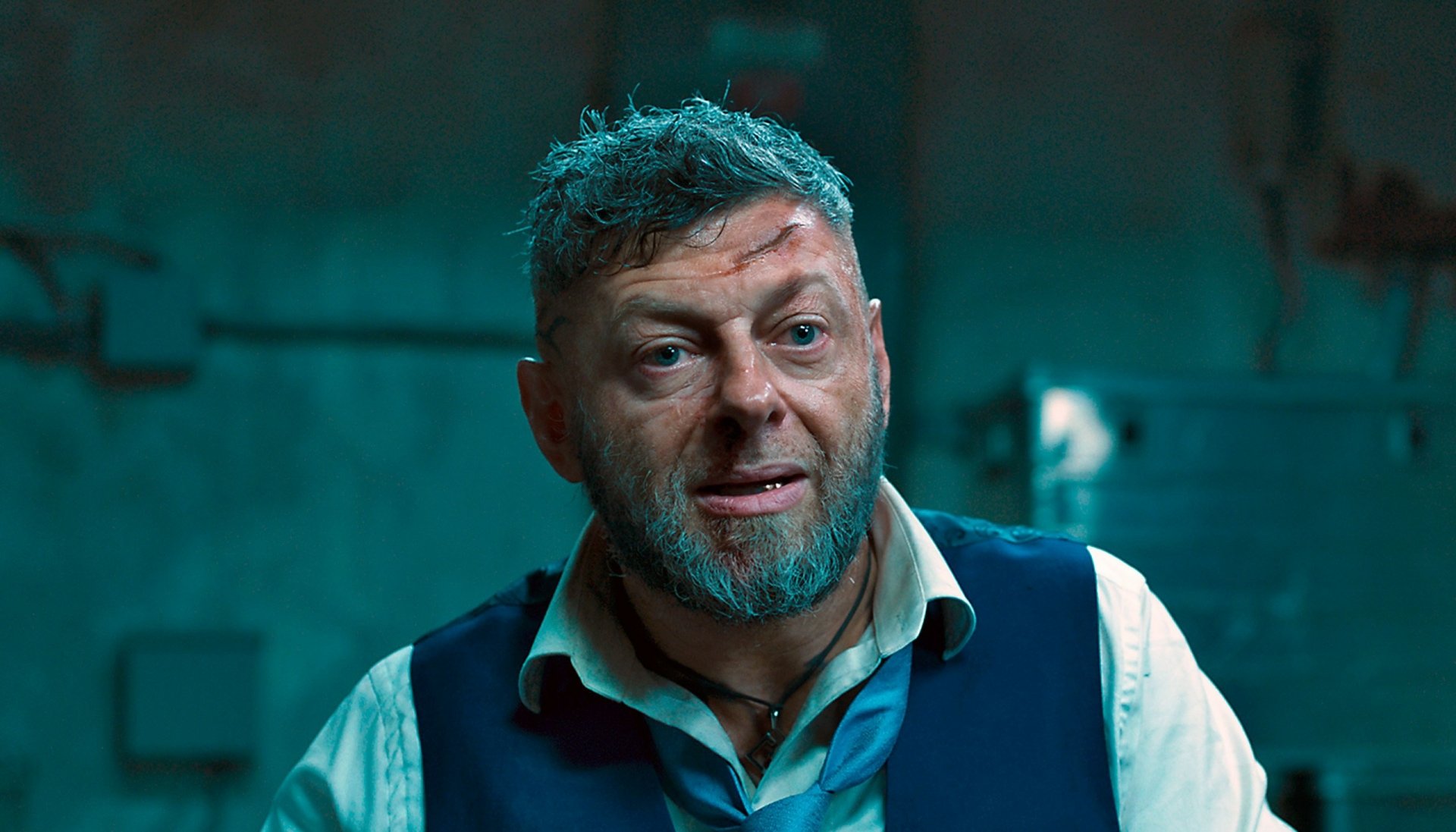 HD desktop wallpaper image from the movie Black Panther, featuring a close-up of a character with short, messy gray hair and a beard, wearing a white shirt and blue vest, in a dimly lit room.
