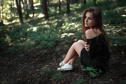 A brunette woman with brown eyes and a black dress sits thoughtfully in a forest, surrounded by blurred greenery and soft bokeh, captured in HD depth of field.